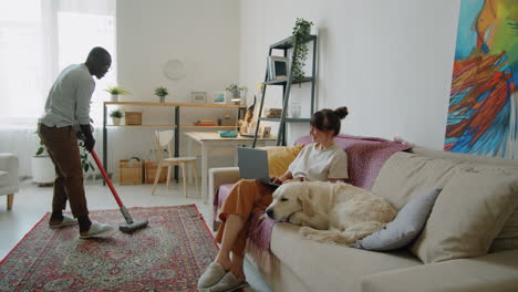 black man vacuuming floor while wife and dog resting on sofa