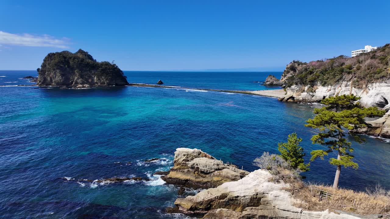 Uma panorâmica ampla em ângulo alto pelas águas cristalinas e turquesa de Dogashima, Japão. A imagem captura os penhascos vulcânicos acidentados, um pinheiro solitário e as icônicas Ilhas Sanshiro.