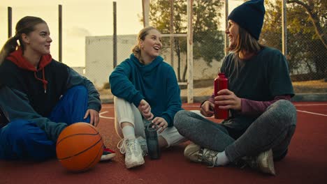 Close-up-a-trio-of-blonde-girls-in-sports-uniforms-hold-sports-bottles-in-their-hands,-sit-on-the-red-floor-on-a-street-court-near-an-orange-basketball-ball,-communicate-and-warm-up-in-the-morning