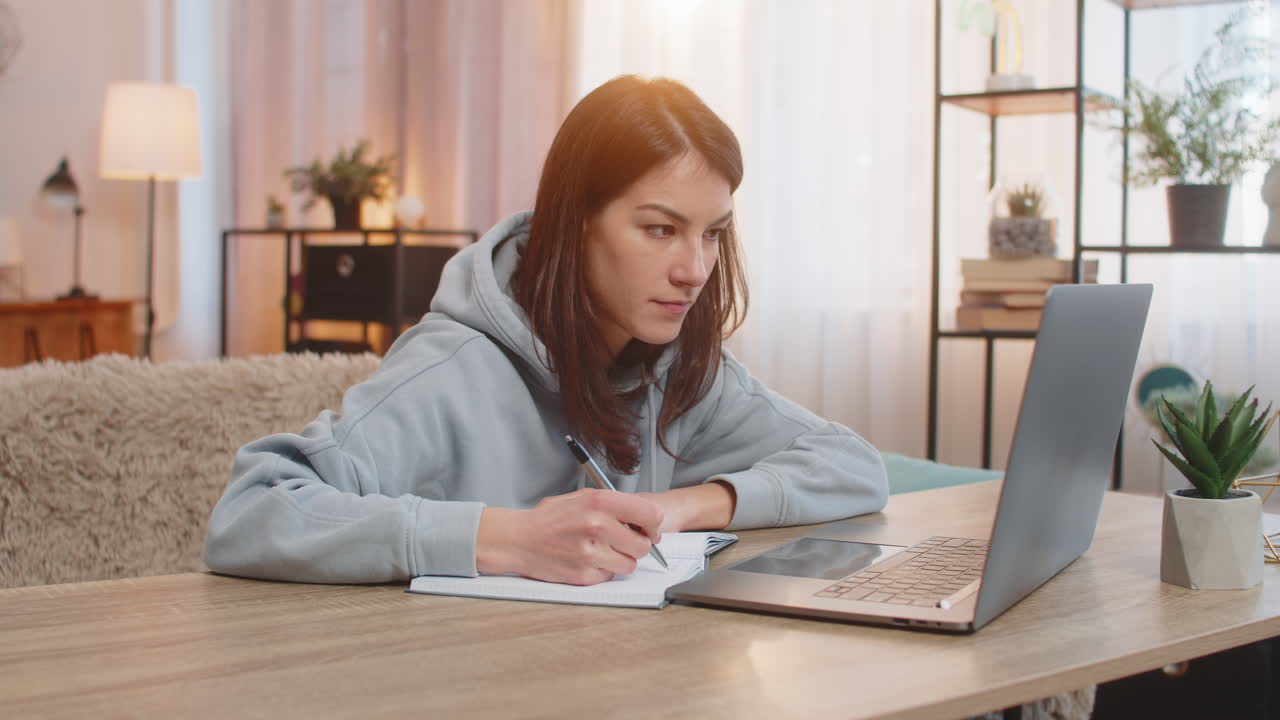 Young woman watching online course on laptop sitting at table on sofa couch taking notes in notebook
