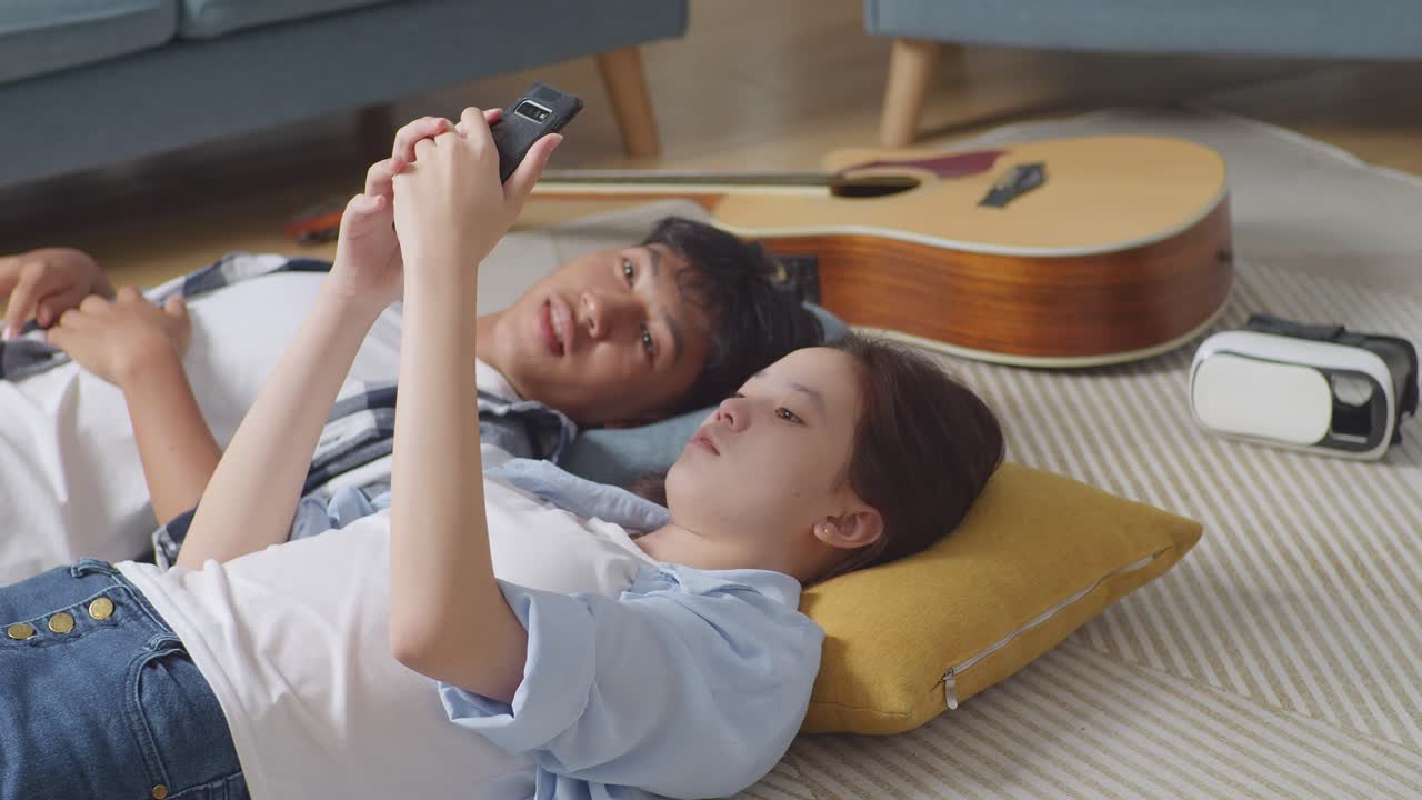 집에서 바닥에 카에 누워있는 십대 아시아 커플: 화가 난 소녀가 스마트폰을 사용하고 남자친구를 무시하고, 화가 난 남자친구가 그녀에게 관심을 기울이려고 시도 한 후 그릴 친구를 혼자 떠난다.