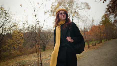 woman with yellow muffler and beret walks through forest, adjusting glasses while holding bag strap, surrounded by autumn foliage