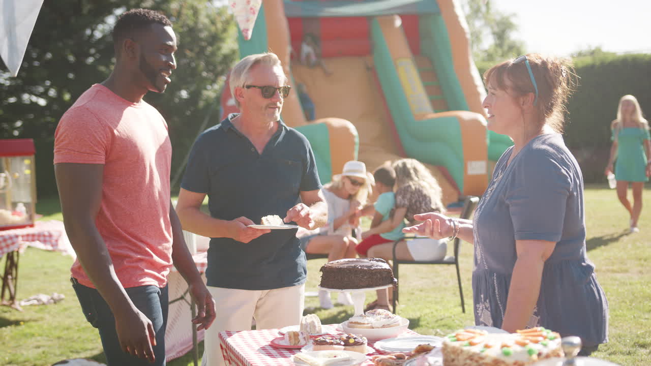 Slow Motion Shot Of Mature Woman Serving On Cake Stall At Busy Summer Garden Fete