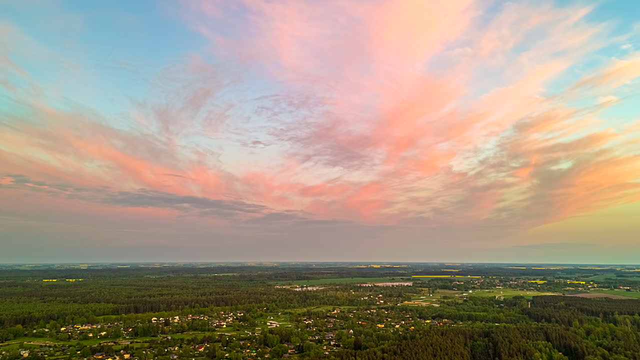 A scenic time-lapse captures a serene sunset painting the vast sky with soft pastel pink and orange clouds above a sprawling rural landscape of green forests and small villages in Latvia