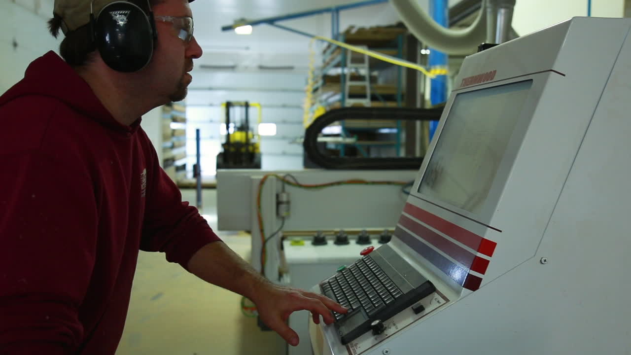 Wide shot of CNC Machine cutting through Melamine Wood. Operator checks up on his calculations at the Computer Station.