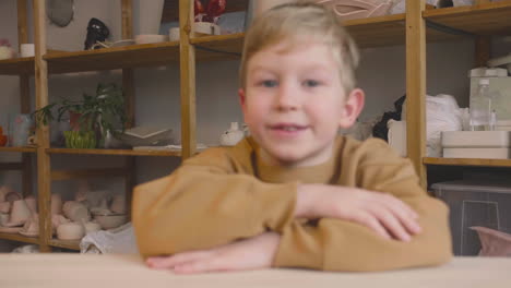 blond kid sitting at a table looking at the camera in a craft workshop