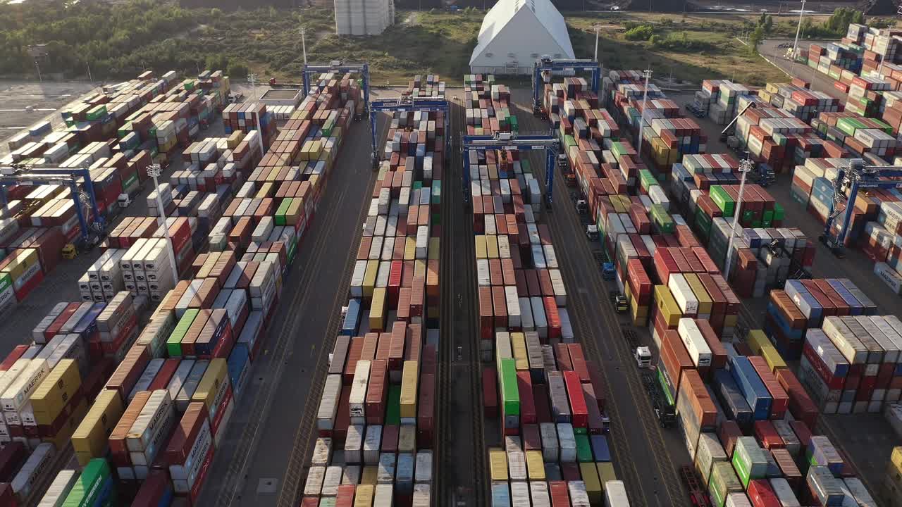 Aerial View Of Gantry Cranes, Shipping Containers, And Logistic Truck Services At Cargo Terminal