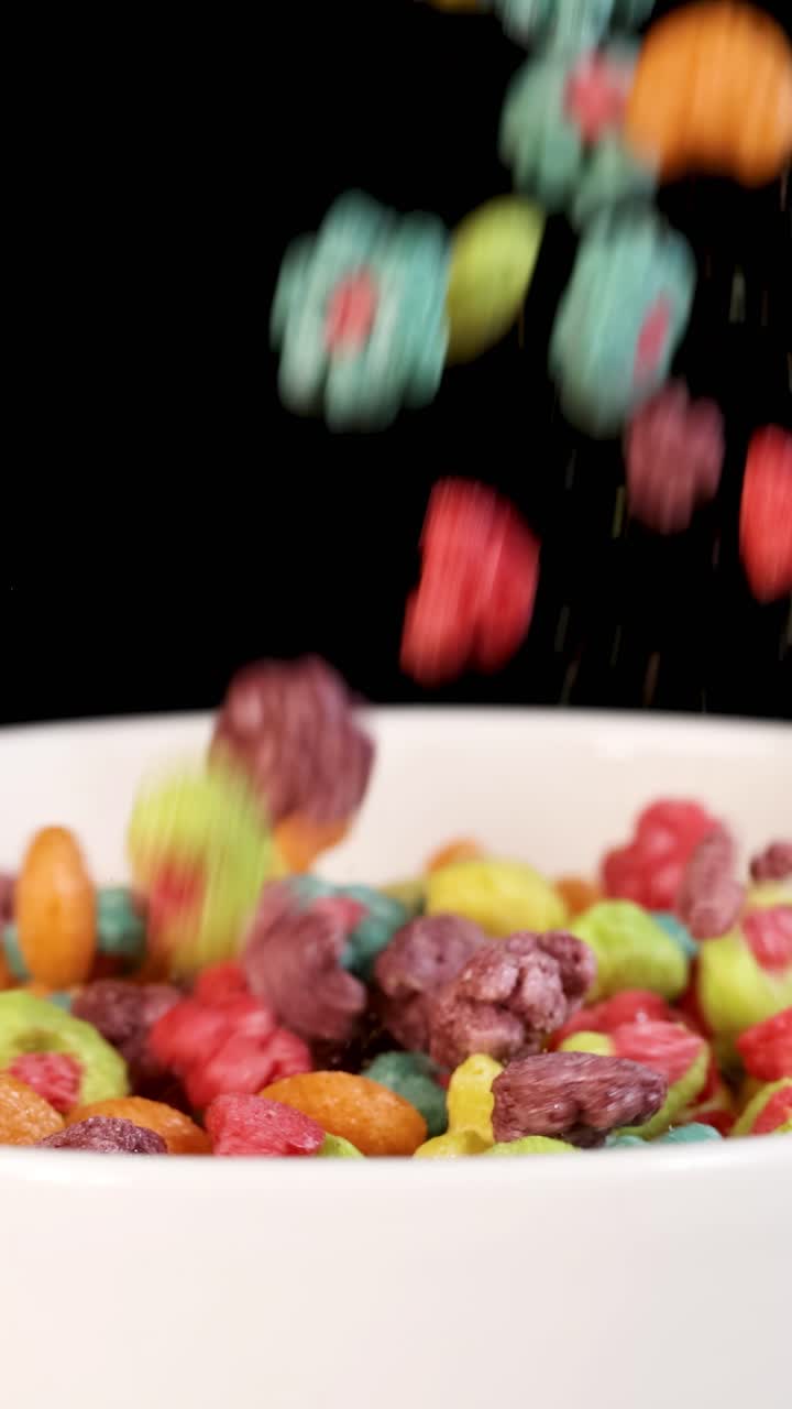 Pezzi di cereali vivaci cadono a cascata in una ciotola bianca su uno sfondo nero, creando una scena dinamica e colorata.