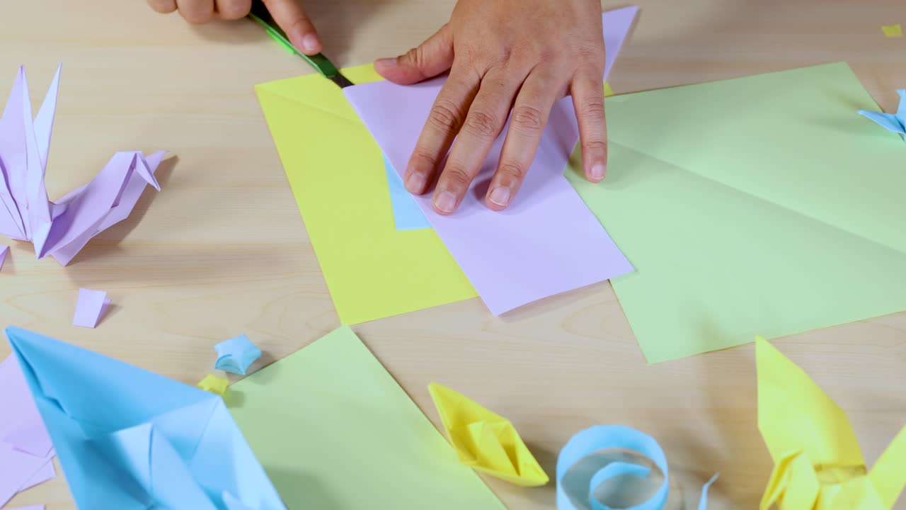 Hands use craft knife to cut pastel paper on desk with origami figures, top-down view