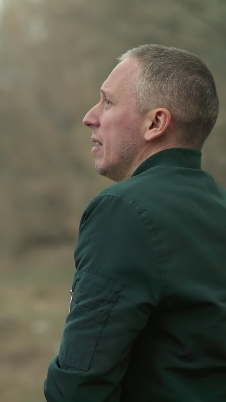 A close view of a man in a green jacket, with a tense expression, reaching into his pocket to draw a pistol while looking over his shoulder in a forested area