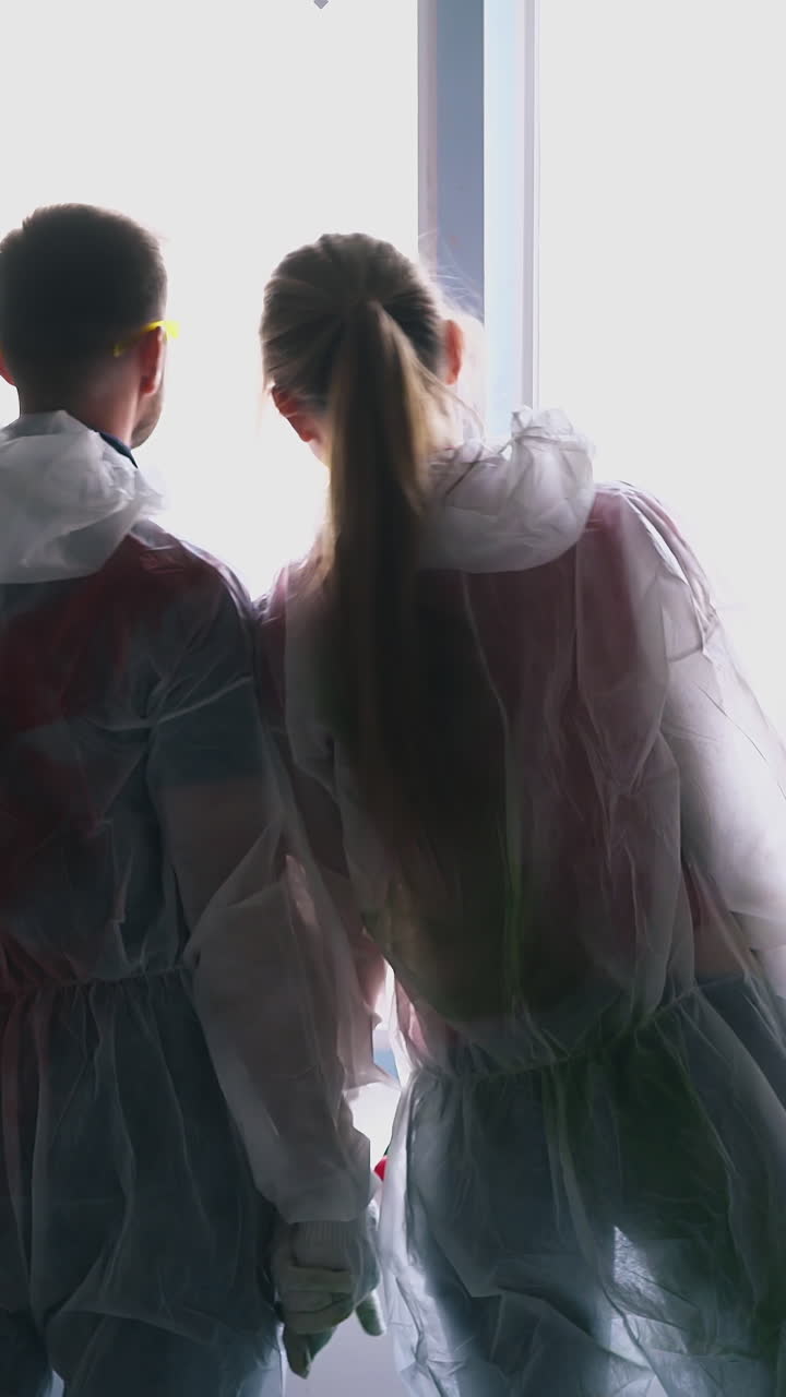 Dolly cam shot: positive young couple wearing protective wear joins hands looking outside window in own apartment