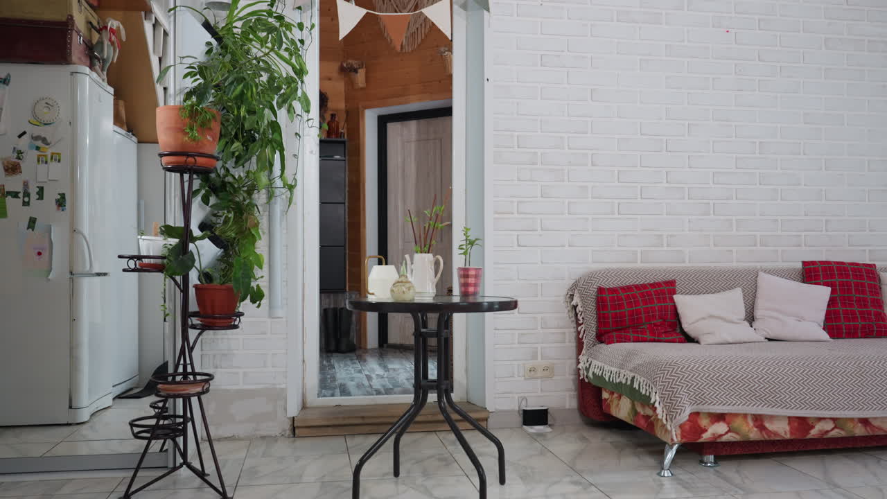 Modern room decorated with green plants on stands next to white brick wall, vintage freezer by open doors, round glass table with pitcher and cup, patterned sofa with cushions