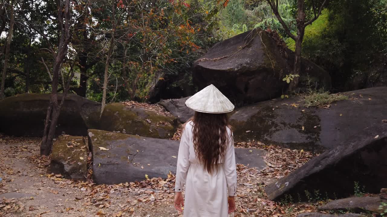 Young european woman traveler in long white dress and vietnamese hat walking in forest near huge boulders, big stones in Vat Phou – ruined Khmer Hindu Temple. Dry leaves, green trees around. Slow motion. Champassak, Laos, Asia.