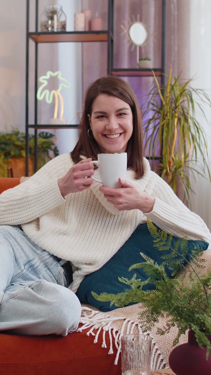 Affascinante giovane donna sorridente che beve una tazza di caffè o tisana sul divano di casa al mattino