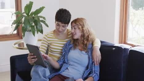 happy caucasian lesbian couple embracing on sofa, using tablet and credit card in sunny house