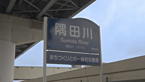 a clear signboard for "sumida river" in both japanese and english, featuring the slogan accessible river beneath an urban overpass.