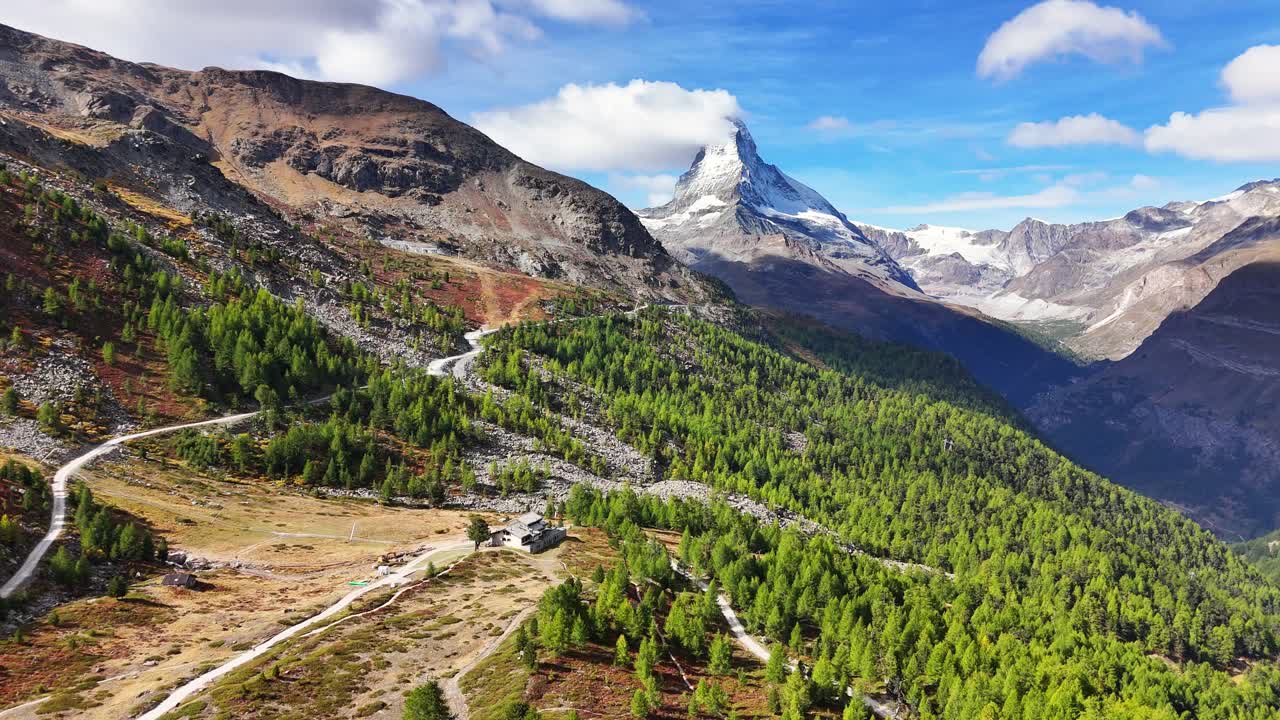 Expansive alpine terrain in Zermatt Riffelalp with the Matterhorn rising above forested valleys, rugged slopes, winding paths and scattered rocky formations beneath a bright blue sky with soft clouds