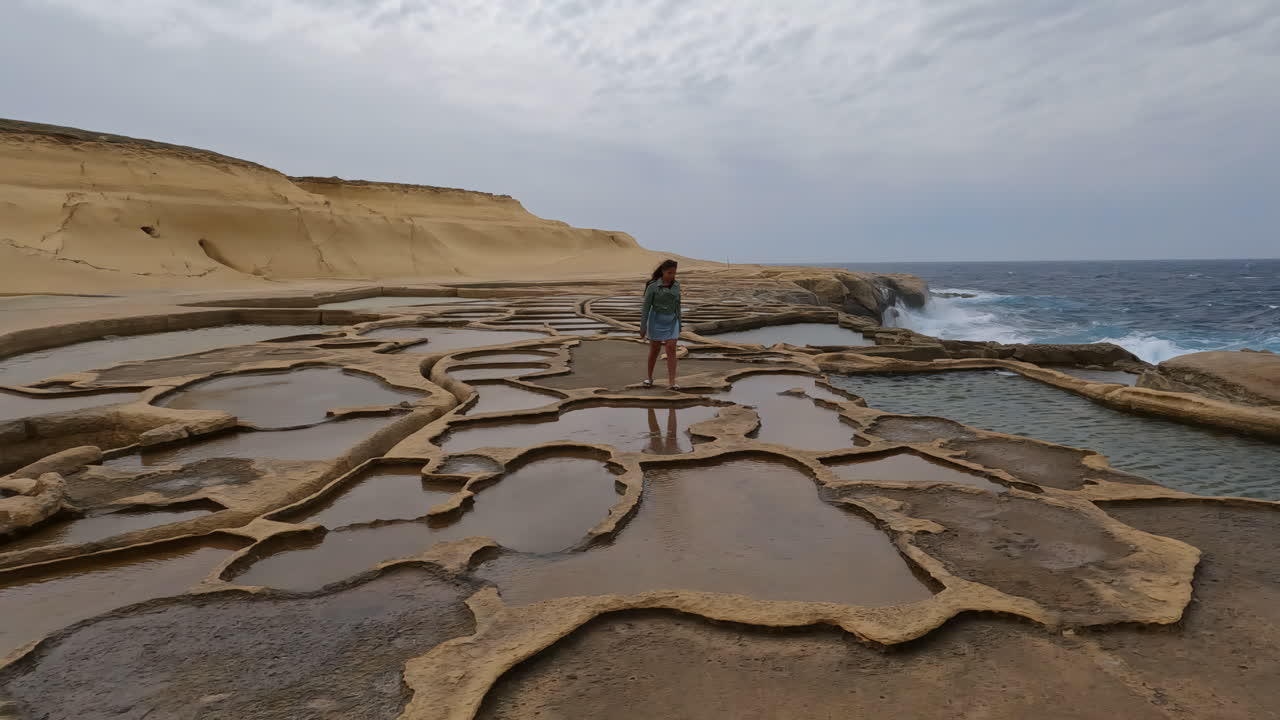 mujer caminando a lo largo de la costa de una playa con piscinas de agua en la piedra erosionada