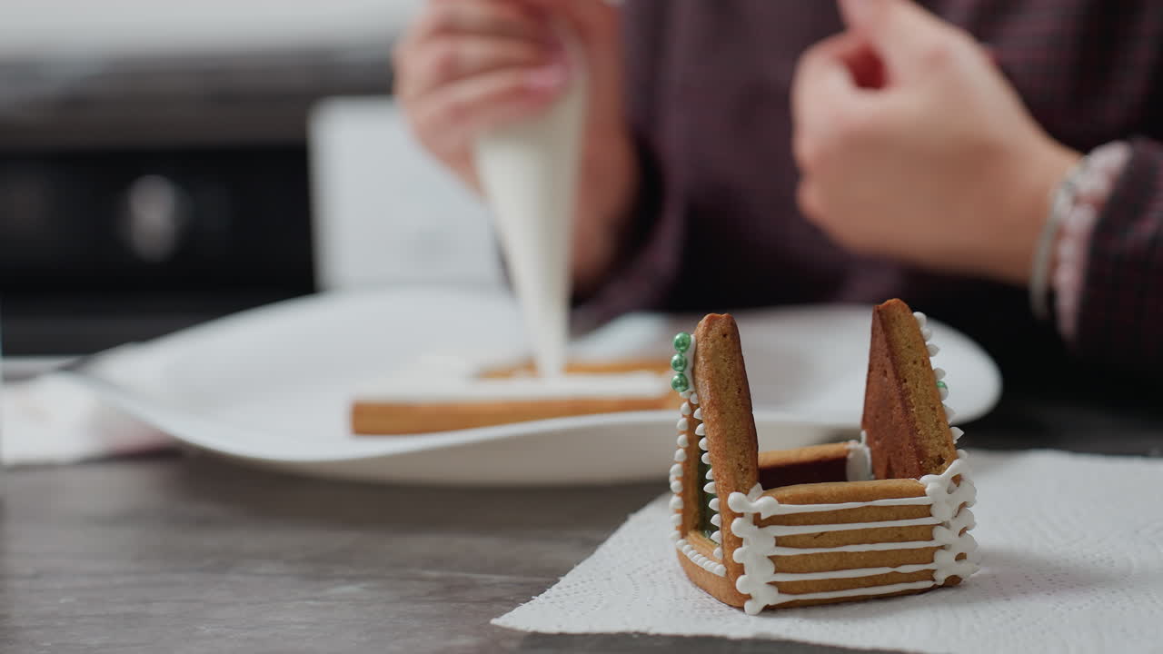 구슬로 된 팔를 가진 여성의 흐릿한 시선 조심스럽게 접시에 아이싱 쿠키, 주방 설정에서 조직에 배치 된  아이싱과 녹색 식용 진주와 함께 장식 된 진저브레드 집