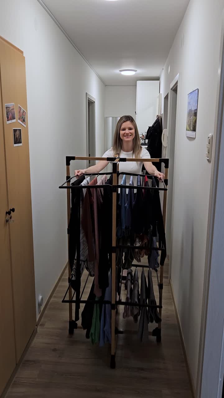 Una donna sorridente spinge uno stendibiancheria portatile pieno di bucato lungo un luminoso corridoio di un appartamento. Ideale per la vita domestica, l'organizzazione della casa e contenuti sulle faccende domestiche.