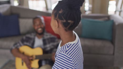 Glückliche-Afroamerikanische-Tochter-Tanzt-Im-Wohnzimmer,-Während-Ihr-Vater-Gitarre-Spielt