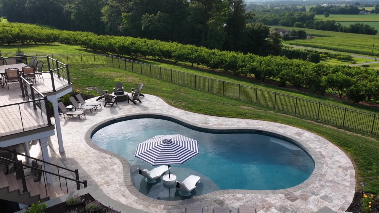 Kidney shaped pool wraps stone patio beside upper deck overlooking orchard rows and open farmland in the United States, wrought iron fence lines luxury backyard property. Aerial drone wide shot