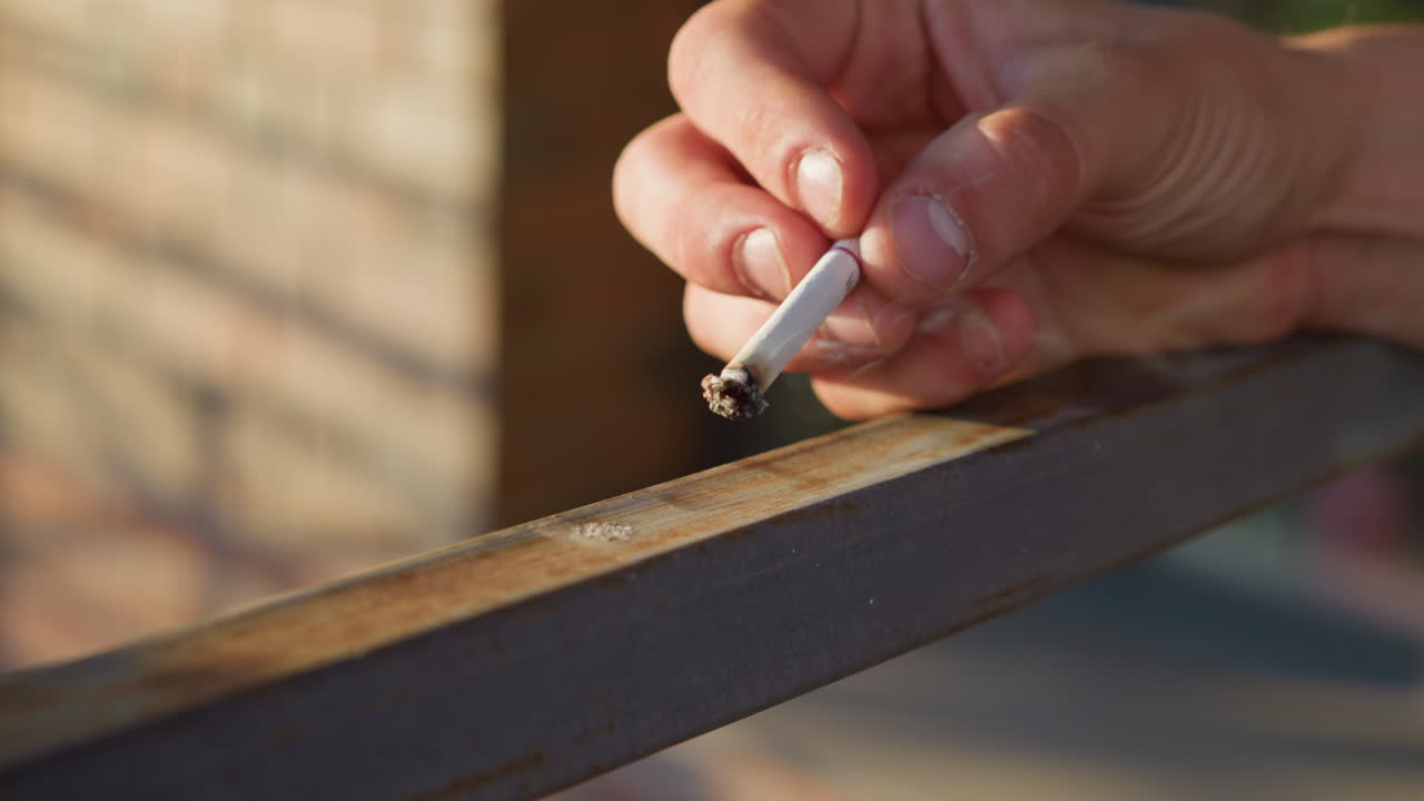hand view of fair skinned person holding cigarette and resting it gently on rusty iron bar under sunlight with shadows falling across surface while smoke subtly escapes from cigarette tip