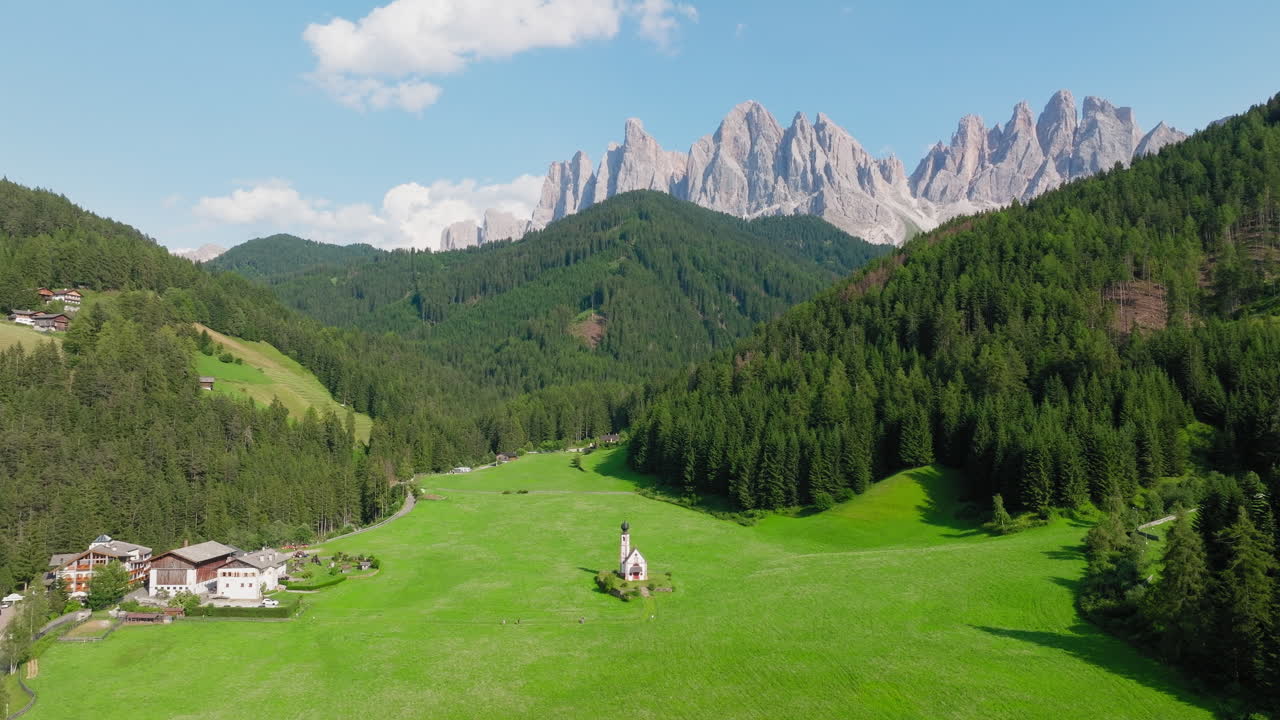 Slow panning drone shot of the iconic Church of St. John in Val di Funes, captured in spring with vibrant green fields and the towering Dolomites in the background
