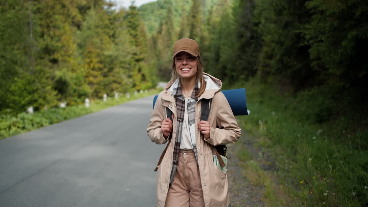 A blonde girl in special clothes for a hiker of light brown color poses near the road being in a mountain forest