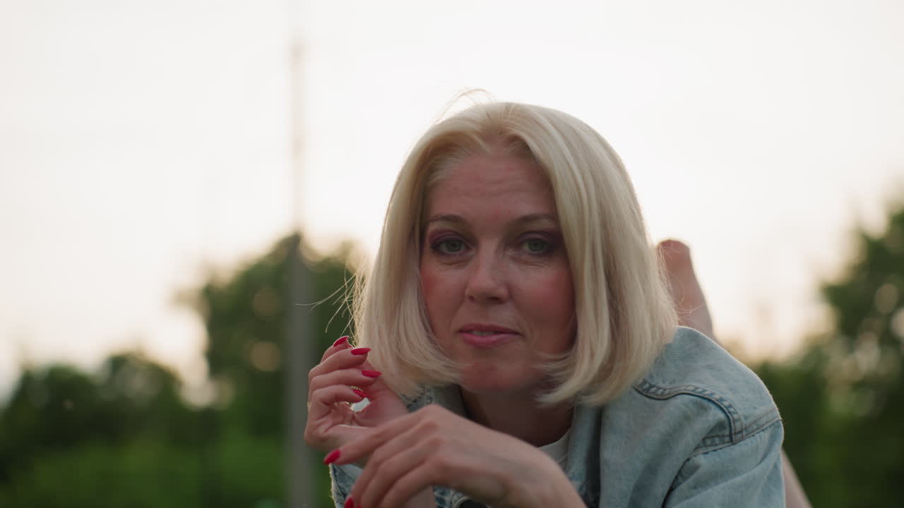 lady with red manicured nails fixing hair while lying on blanket in park using hands near head, blurred people playing in background, golden evening light, relaxed peaceful mood