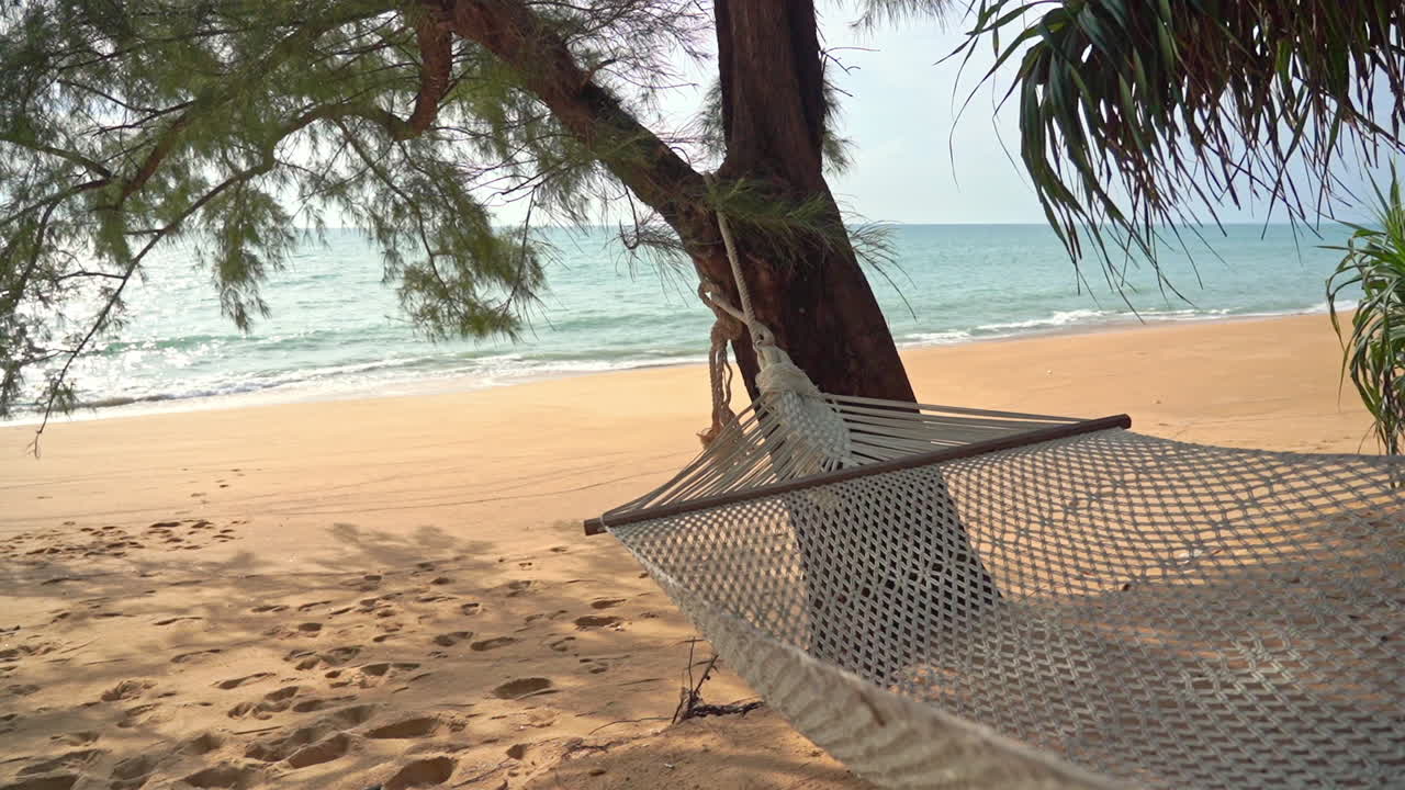 hamaca a la sombra de un árbol junto a una playa tropical de arena y olas marinas ligeras, fotograma completo a cámara lenta