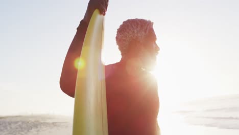 senior african american man holding surfboard on sunny beach