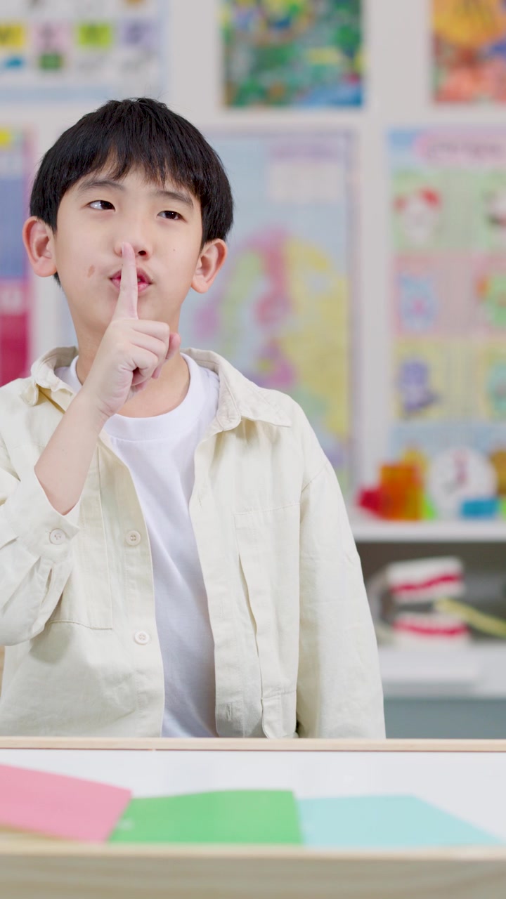 Murid laki-laki Asia yang tersenyum memberi isyarat untuk tenang di ruang kelas yang terang, bidikan medium statis, pencahayaan lembut