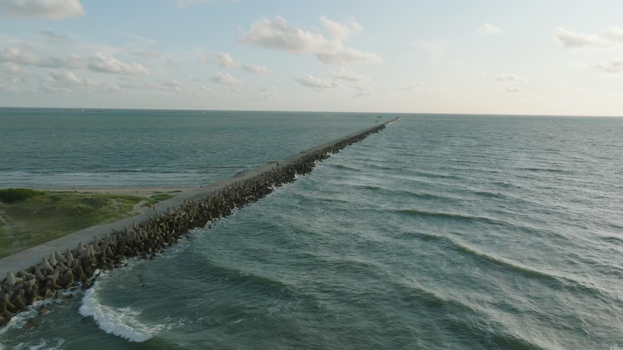 Vista aérea panorámica del muelle de hormigón del Puerto de Liepaja, costa del mar Báltico, tarde de verano soleada, luz de la hora dorada, grandes olas rompiendo, toma amplia de dron avanzando.