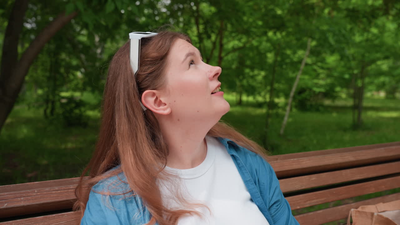 Joven sentada en un banco del parque bebiendo agua de una botella de plástico en un caluroso día de verano, refrescante momento bajo la brillante luz del sol, rodeada de árboles verdes.