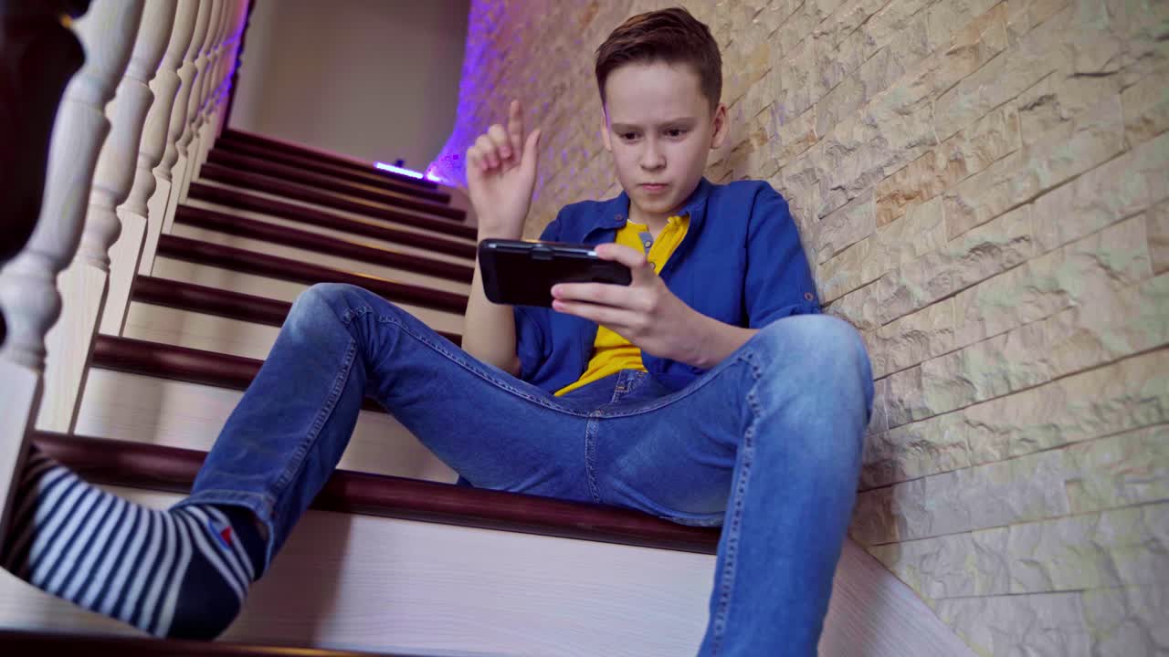 View from below on a boy sitting on stairs at home. Happy teenager playing on his mobile phone and wins the game. Gaming concept.