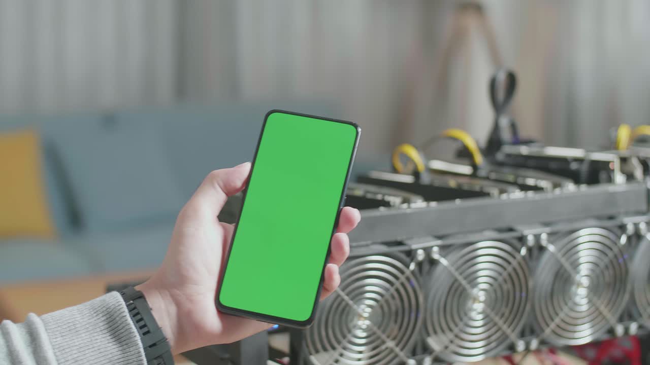 cerca de la mano del hombre sosteniendo un teléfono inteligente de pantalla verde frente a la plataforma de minería para la minería de criptomonedas