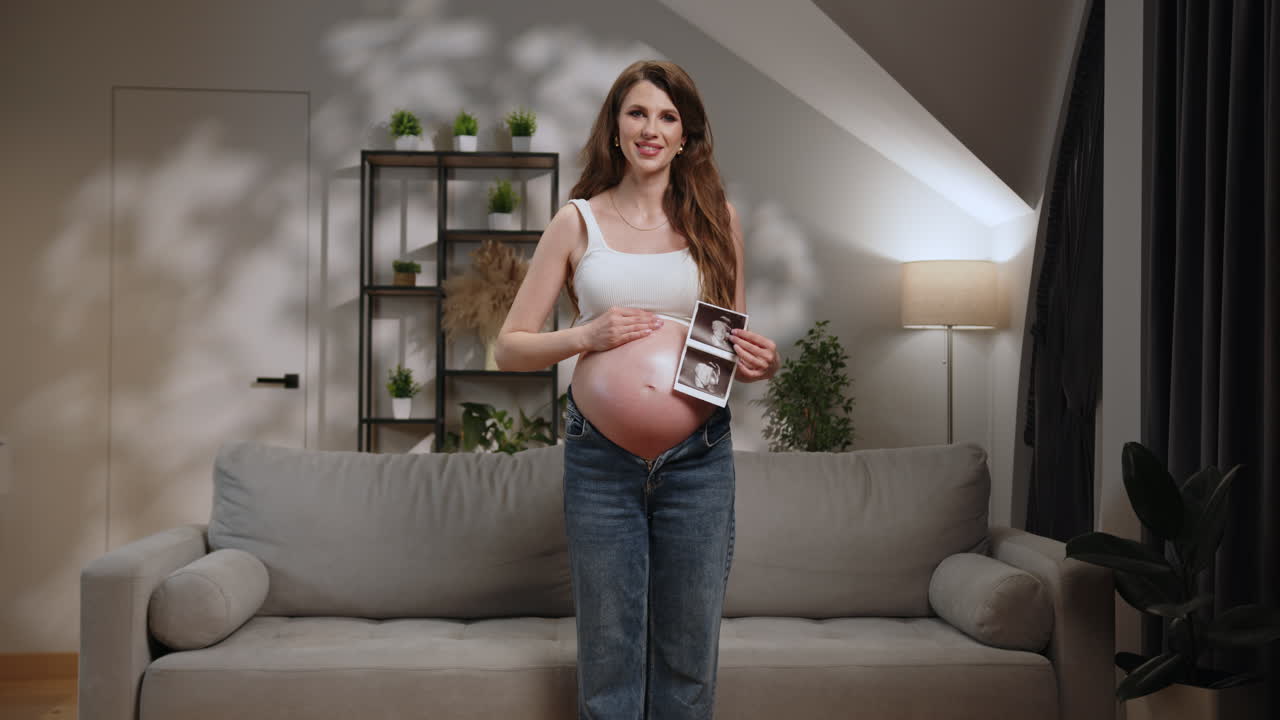 Smiling pregnant woman in casual clothes stands in living room holding ultrasound images of her baby