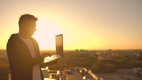 a male freelance programmer sits on a skyscraper roof with a laptop and beer typing code on a keyboard during sunset. remote work