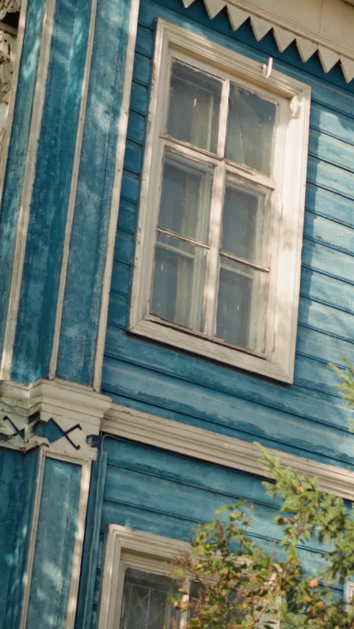 Casa de madera azul con ventana iluminada por el sol, adornos ornamentados, cortina de encaje y pintura desgastada. El revestimiento texturizado y las sombras suaves crean un ambiente acogedor y nostálgico alrededor del exterior de una casa de campo histórica enmarcada por un jardín.