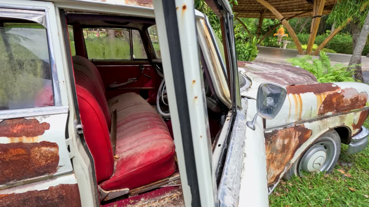 Person closes door of weathered antique car with red interior, natural daylight, rural outdoor setting