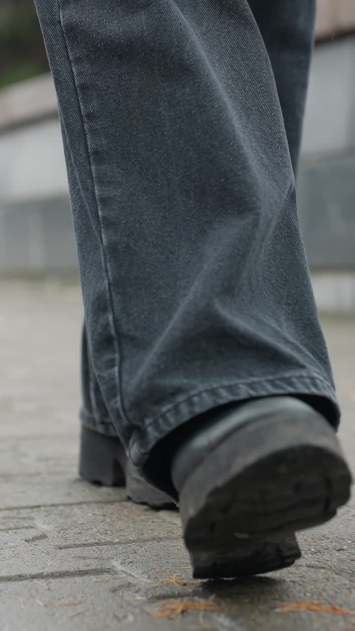 Close up of someone's black trousers and black boots walking slowly toward camera on wet paved urban path, face not visible, capturing motion of steps and rainy atmosphere in quiet autumn setting
