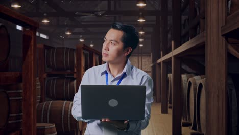 asian male professional worker standing with his laptop in cellar of winery, he observes by looking around before he come to concentrating with his laptop
