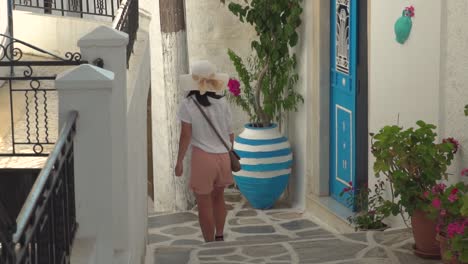 Mädchen-Im-Sommerlichen-Wanderoutfit-Läuft-Durch-Die-Typisch-Enge-Griechische-Gasse-Mit-Blauen-Türen-Und-Pflanzen