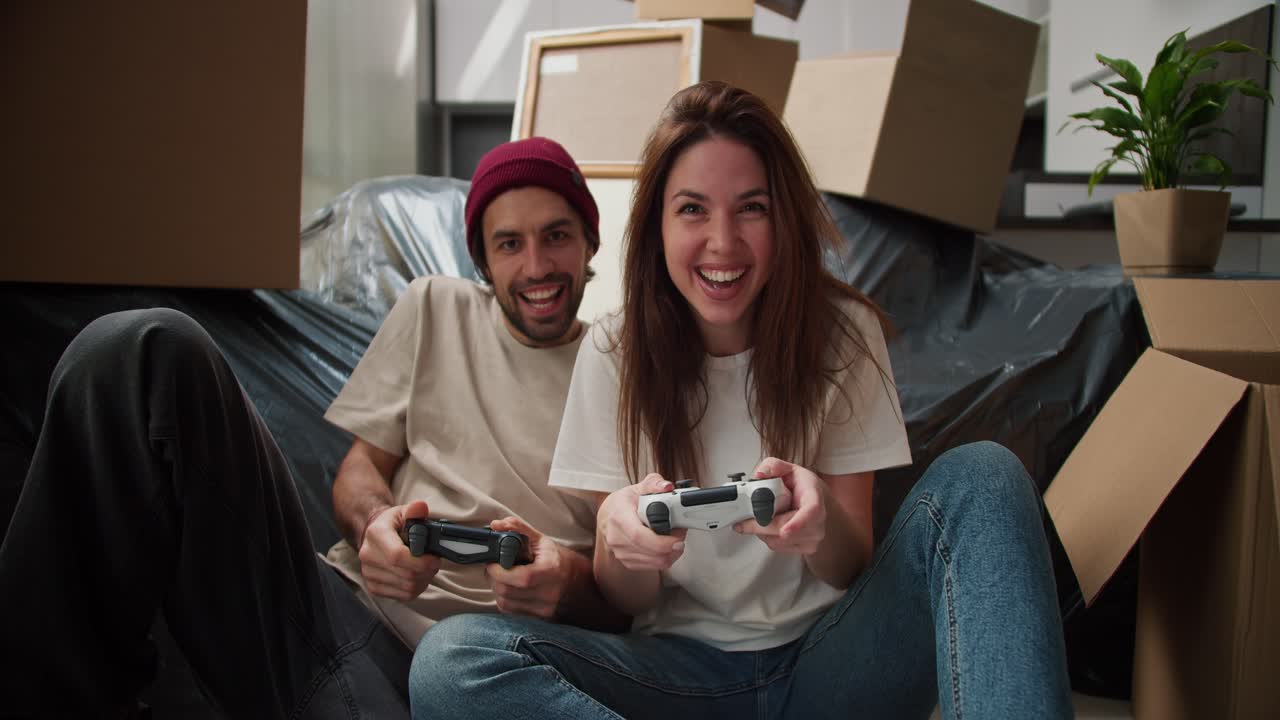 Happy brunette girl in a white T-shirt plays video games with her boyfriend using joysticks has a lot of fun sitting on the floor near the sofa packed in black plastic and a lot of boxes around after moving