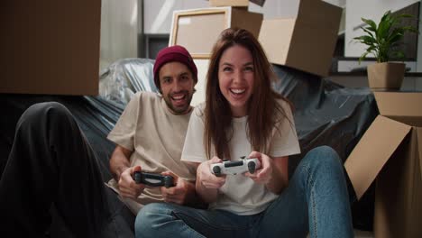 Una-Chica-Morena-Feliz-Con-Una-Camiseta-Blanca-Juega-Videojuegos-Con-Su-Novio-Usando-Joysticks-Y-Se-Divierte-Mucho-Sentada-En-El-Suelo-Cerca-Del-Sofá,-Envuelto-En-Plástico-Negro-Y-Con-Muchas-Cajas-Alrededor-Después-De-Mudarse.