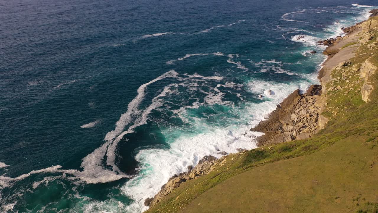 vuelo con un dron en un acantilado en el mar de cantabria dejando un prado verde viendo las olas chocar contra las rocas con una variedad de colores azules en el mar en una tarde de verano en cantabria españa