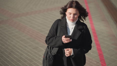 woman walking outdoors, looking around briefly before focusing on her phone, with her hair slightly blowing in the wind, she wears a black coat, carries a bag, and walks on a patterned urban pathway