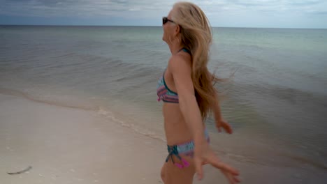 mujer madura rubia juguetona con gafas de sol y bikini corriendo por la playa y mirando hacia atrás a la cámara