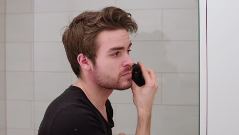 young male man trimming and shaving his beard in front of mirror in bathroom